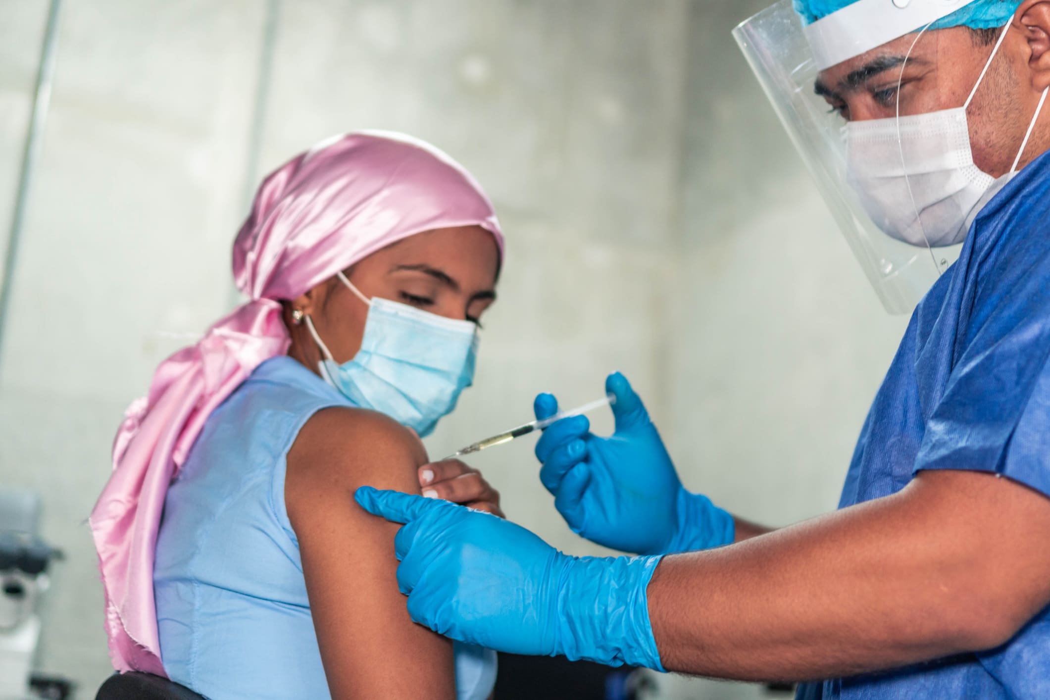 Mujer con cáncer usando pañuelo rosa y mascarilla recibiendo una vacuna administrada por un profesional sanitario con equipo de protección.