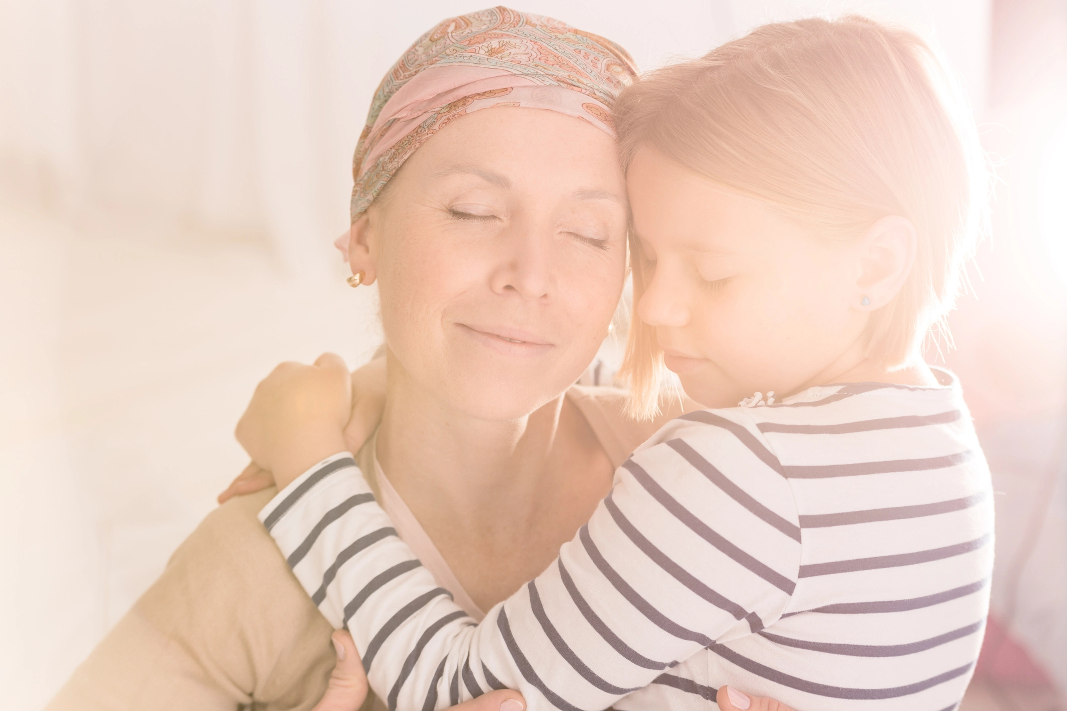 Madre con cáncer abrazando con ternura a su hija pequeña, ambas con los ojos cerrados en un momento de conexión emocional.