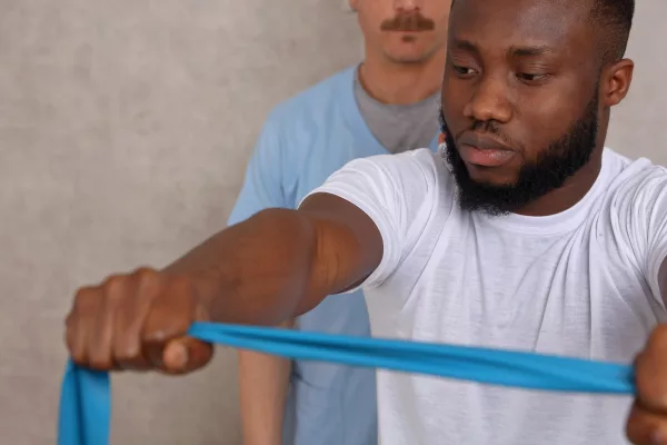 Un hombre con camiseta blanca está realizando ejercicios de rehabilitación con una banda elástica azul para la recuperación de tendinitis en el hombro, guiado por un fisioterapeuta con camiseta azul y bigote, que se encuentra detrás de él, en un ambiente clínico con paredes de color neutro
